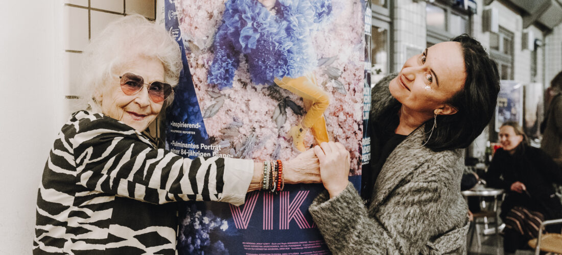 Vika und Regisseurin Filmproduzentin Agnieszka Zwiefka umarmen mit verschmitzten Gesichern eine Säule, auf dem ein Plakat ihres Films prang